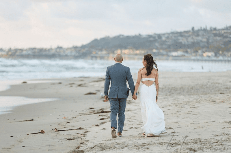 A true-to-life color rear view photo of a bride and groom holding hands as they walk on a beach.