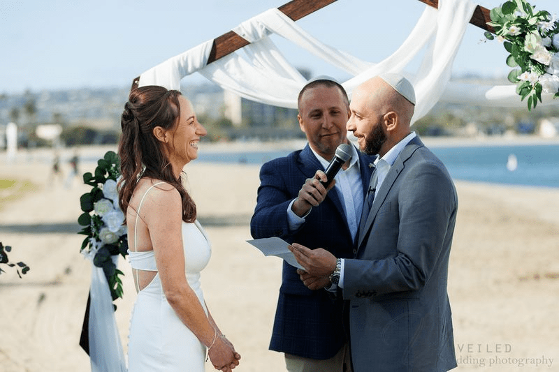 True-to-life color beach wedding ceremony with bride and groom exchanging vows under arch by ocean