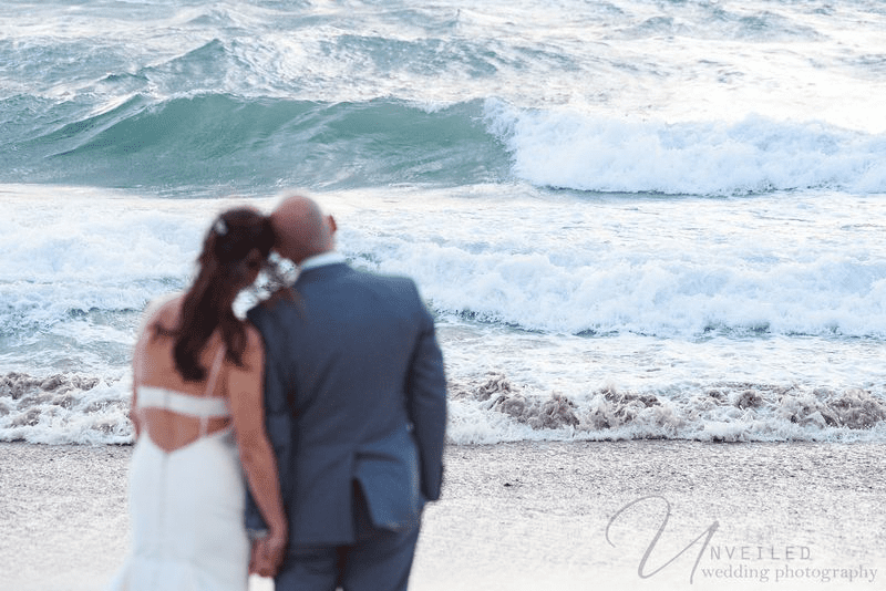 A True-to-life color back view of a couple holding hands by the beach as they watch the waves.