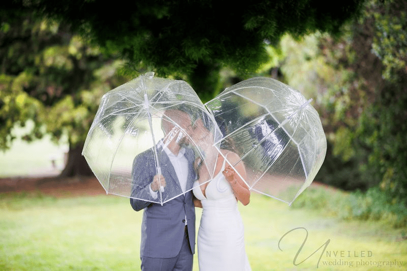 A bride and groom in a True-to-life color photograph are kissing under umbrellas in a park.