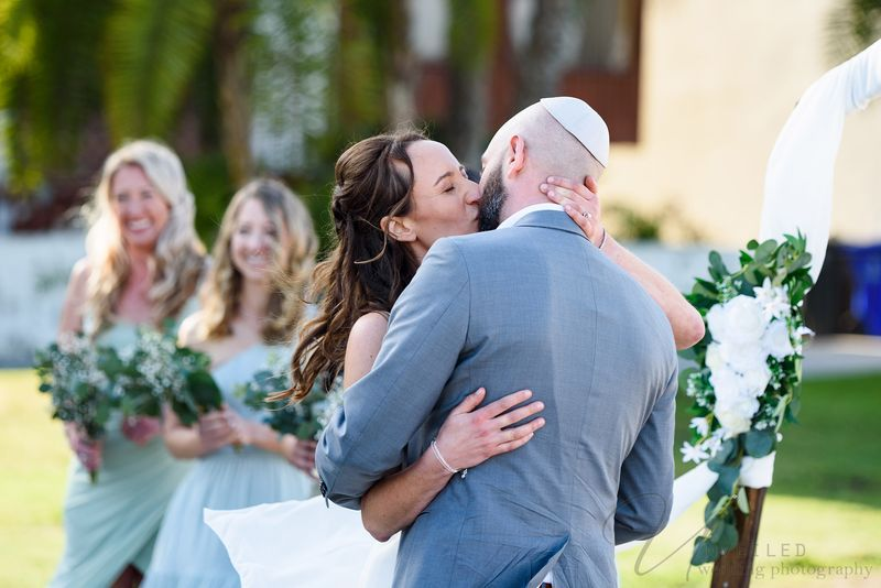 True-to-life color bride and groom share a kiss at outdoor wedding ceremony with bridesmaids smiling behind