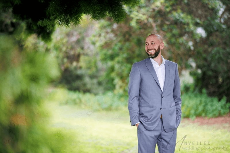 A true-to-life color photograph of a joyful groom in a grey suit, smiling with hands in his pockets.