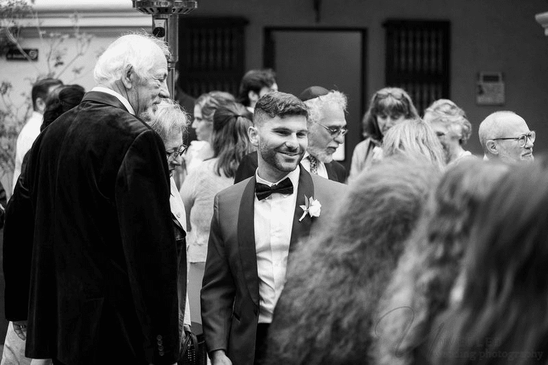 Black and white photo of a smiling groom in a tuxedo surrounded by guests at a wedding reception.