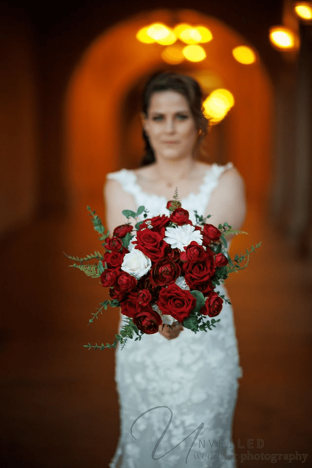 A bokeh photograph shows a blurred bride presenting a sharp red and white rose bouquet to the camera.