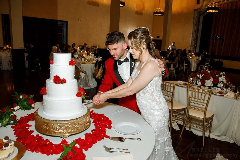 A newlywed couple cuts their four-tier white wedding cake decorated with red roses at their reception.