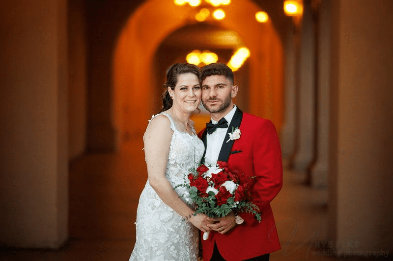 
A bride and groom pose together, with the groom in a red jacket and the bride holding red roses.