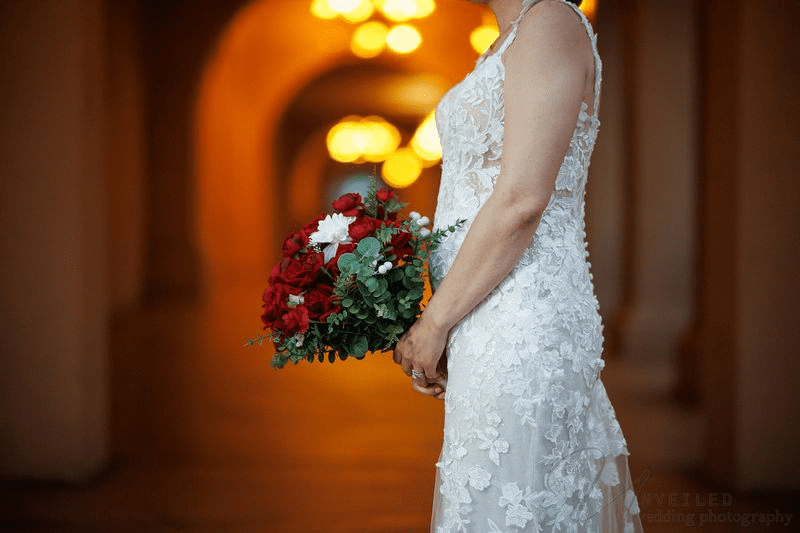 A bride in a lace dress holds a bouquet of red and white roses against a warm, blurred background.