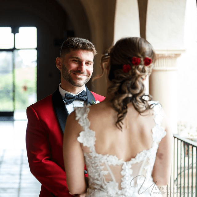A groom in a red jacket smiles at his bride, whose back is to the camera with red flowers in her hair.