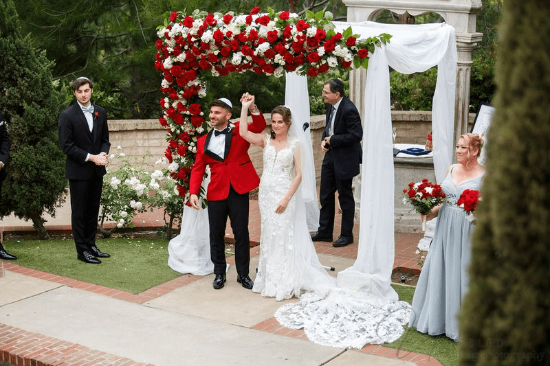 A bride and groom hold hands under a floral chuppah during their outdoor wedding ceremony.