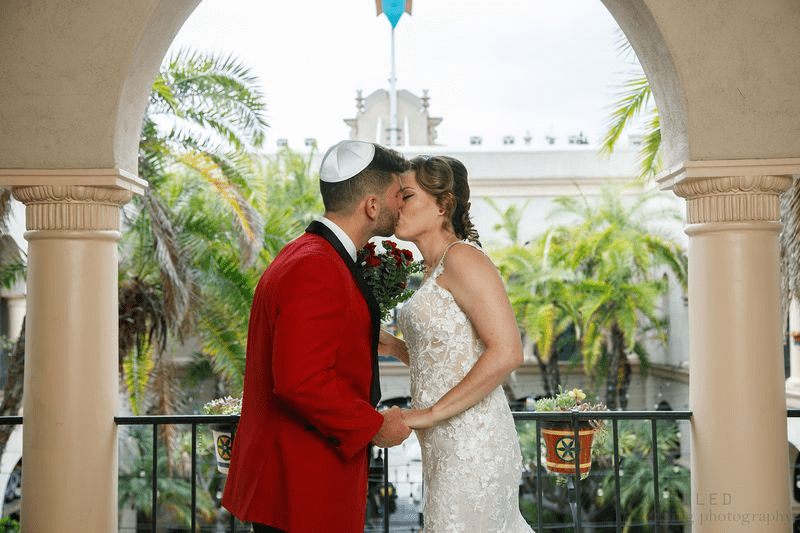 A wedding couple sharing a kiss on a balcony with palm trees and Mediterranean architecture.