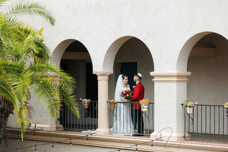 A bride and groom share a moment on a balcony under arches, with the groom in a red jacket.