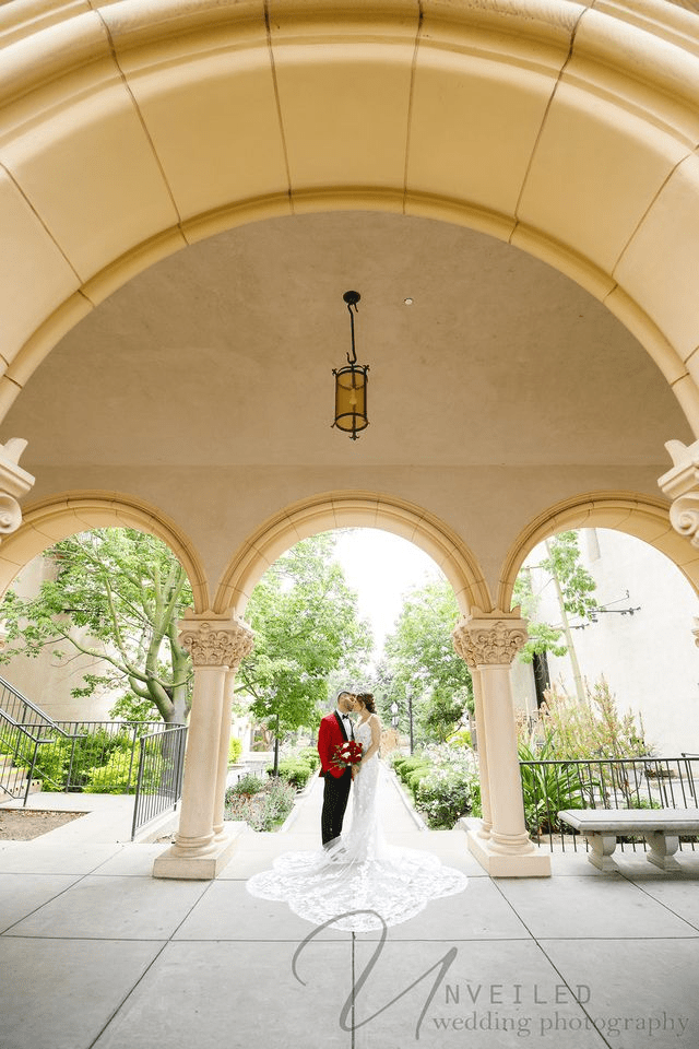 Bride and groom kiss outdoors under a stone archway, framed by elegant columns and garden greenery.