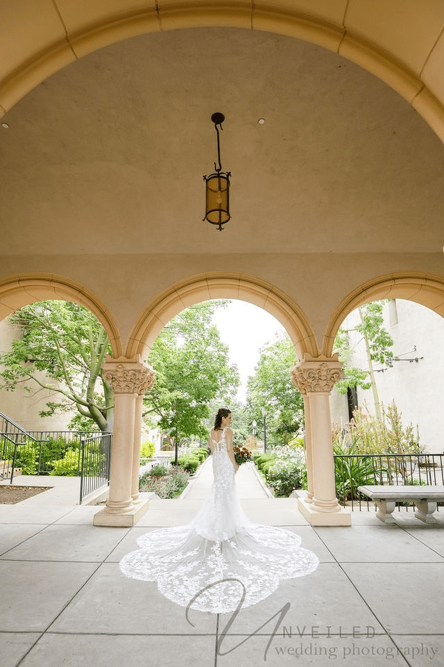 
A bride in a lace gown with a long train stands under a stone archway in a lush garden setting.