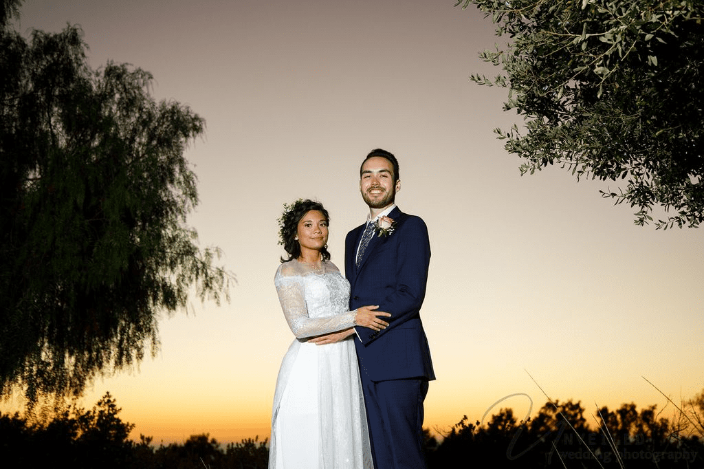 Newlyweds in a white dress and blue suit smile together against a sunset horizon.