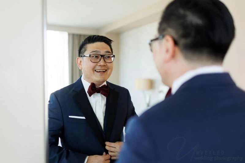
A smiling groom in a navy tuxedo and burgundy bowtie looking at his reflection in a mirror.