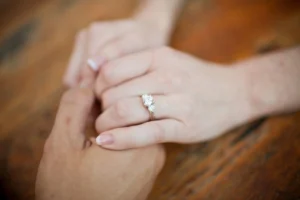 A warm, close-up shot of a couple holding hands outdoors, with the focus on the woman’s hand resting on top, beautifully showcasing her brilliant diamond engagement ring on her finger.