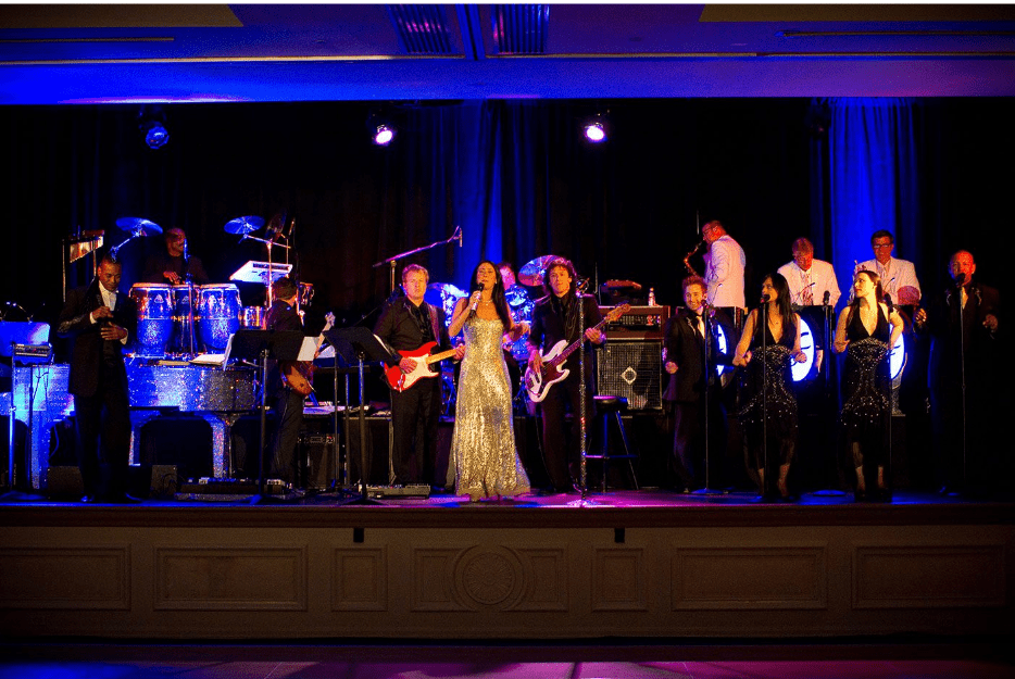 A large live band performing on a stage under vibrant blue and purple lighting.