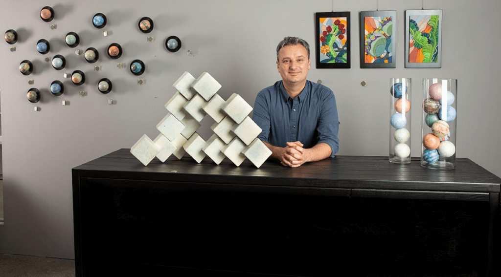 An artist sitting behind a desk with his geometric sculptures and artwork.