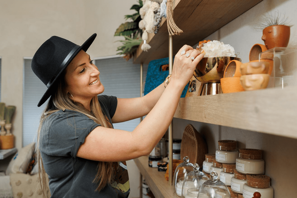A smiling woman in a black hat arranges items on a wooden retail shelf.