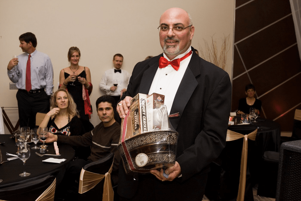 A smiling man in a tuxedo holding a gift basket filled with crackers at a formal event.