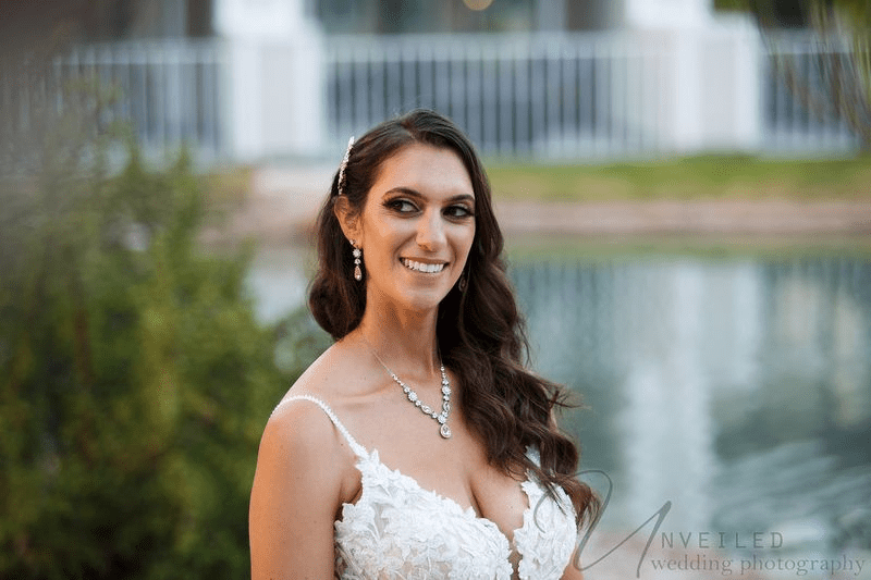 Smiling bride in a white lace gown looking aside with a lake in the background.