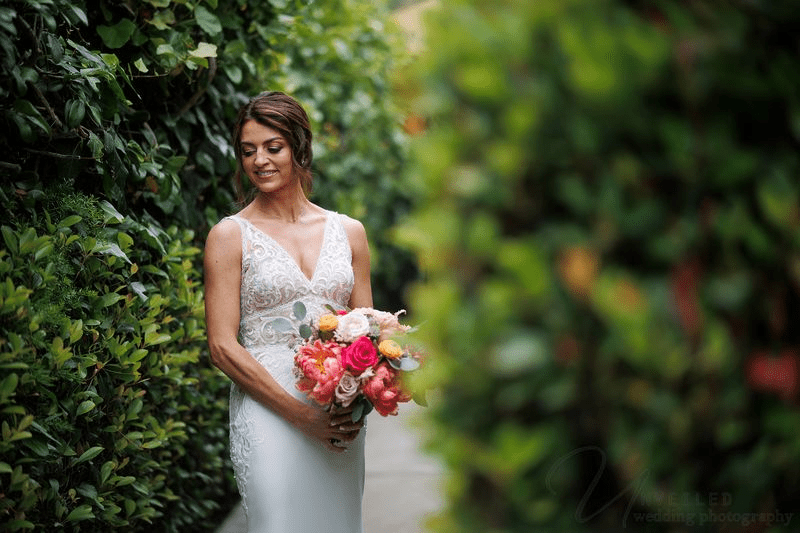 A bride in a lace gown smiles while holding a colorful bouquet against a lush green hedge.