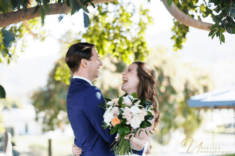 Happy bride and groom laughing together outdoors with a floral bouquet.