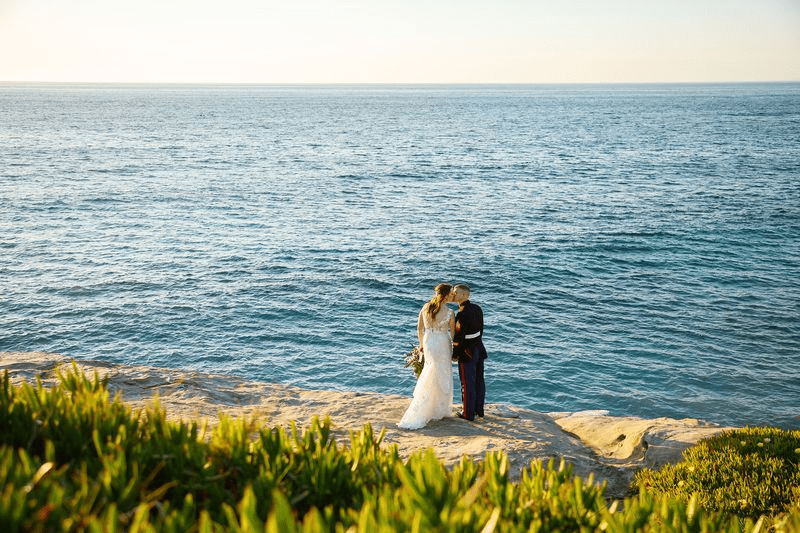 A bride and groom share a close embrace outdoors as the groom kisses the bride's cheek.