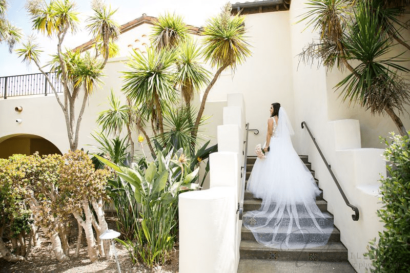 Bride in white gown with long veil walking up outdoor stairs near palm trees.