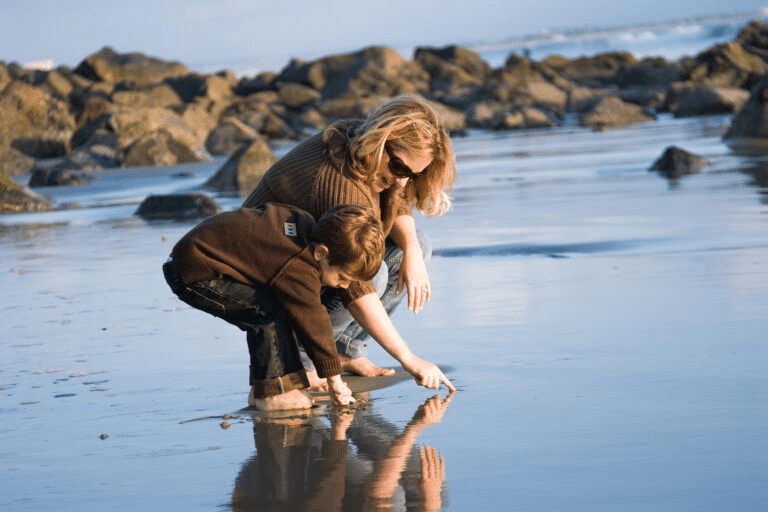 A mother and son squatting on a wet beach, pointing at their reflection.