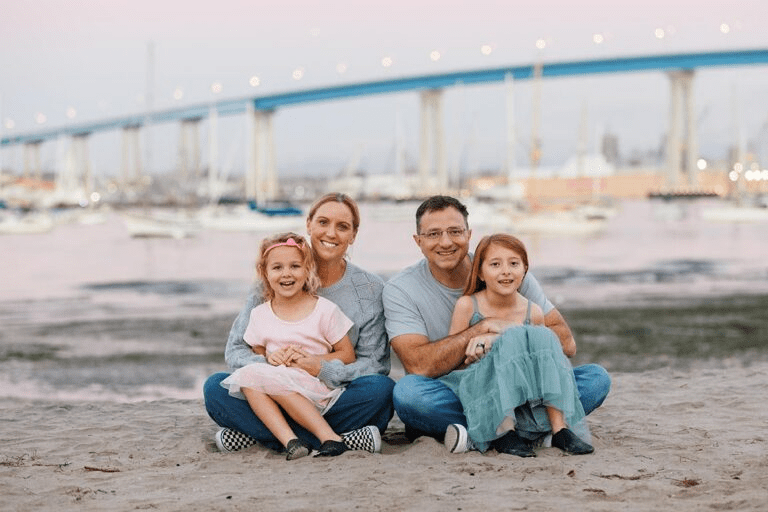 A family of four sitting on a beach with a large bridge in the background.