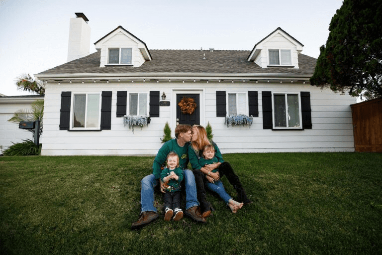 A family sitting on the lawn in front of a white house with black shutters.