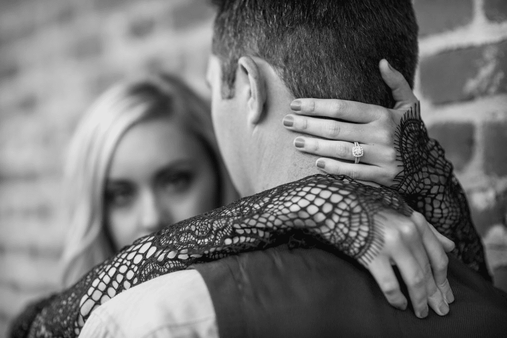 A close-up, black and white photo of a blonde woman smiling softly as she embraces her partner from behind, with her hand resting on the back of his neck, prominently displaying a diamond engagement ring.