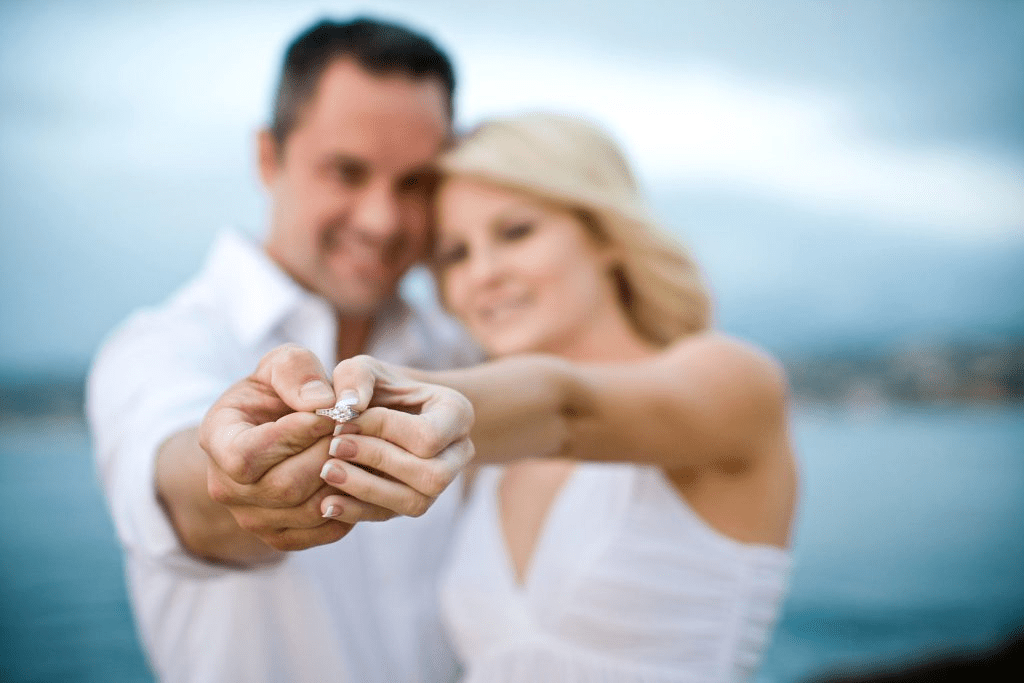 A close-up portrait of a newly engaged couple smiling and embracing outdoors, with the woman looking joyfully at the camera while showing off her new engagement ring.