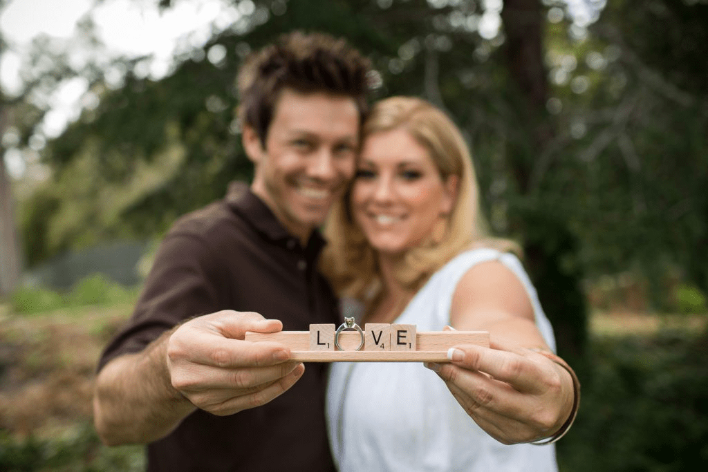 A close-up shot of a newly engaged couple smiling happily in the background, out of focus, while the man holds a wooden Scrabble tile holder in the sharp foreground spelling out the word "LOVE" with a diamond engagement ring acting as the letter "O".