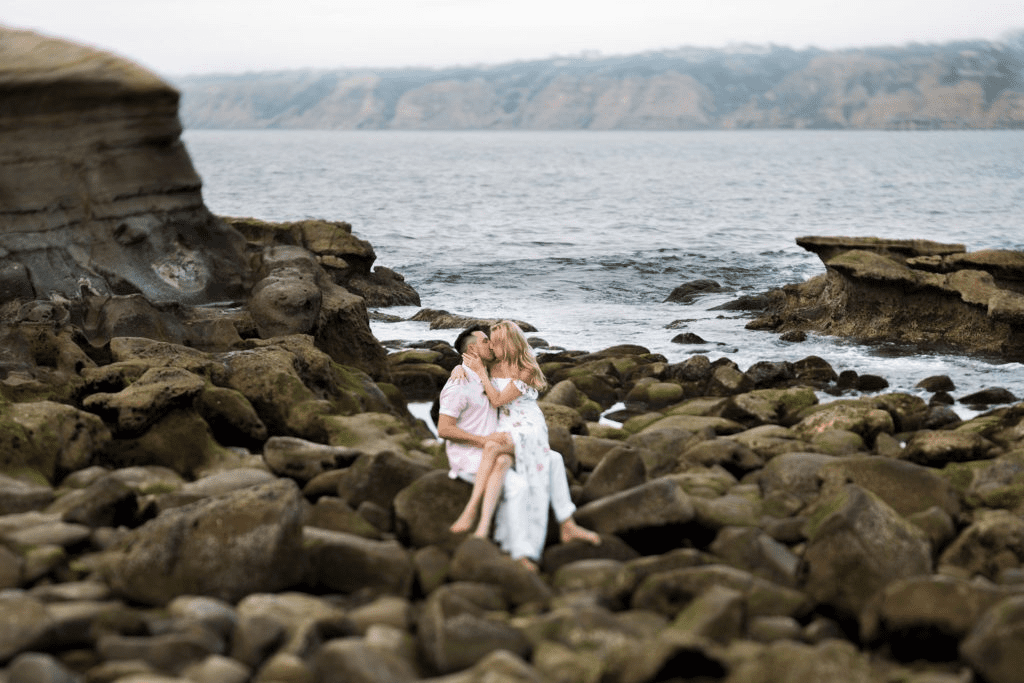 A couple sitting together and embracing on a large, rugged field of coastal boulders and rocks by the ocean tide.