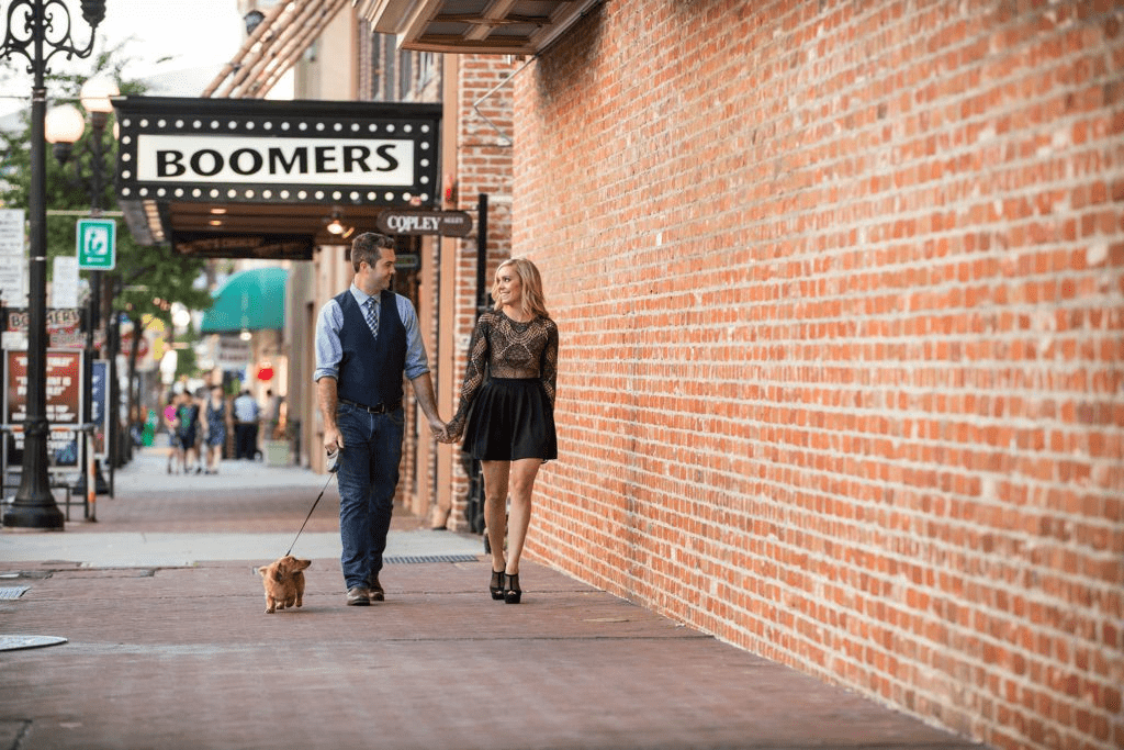 A smiling couple holding hands while walking their dachshund puppy along a brick sidewalk next to a historic brick building in downtown San Diego, with the "BOOMERS" sign visible in the background
