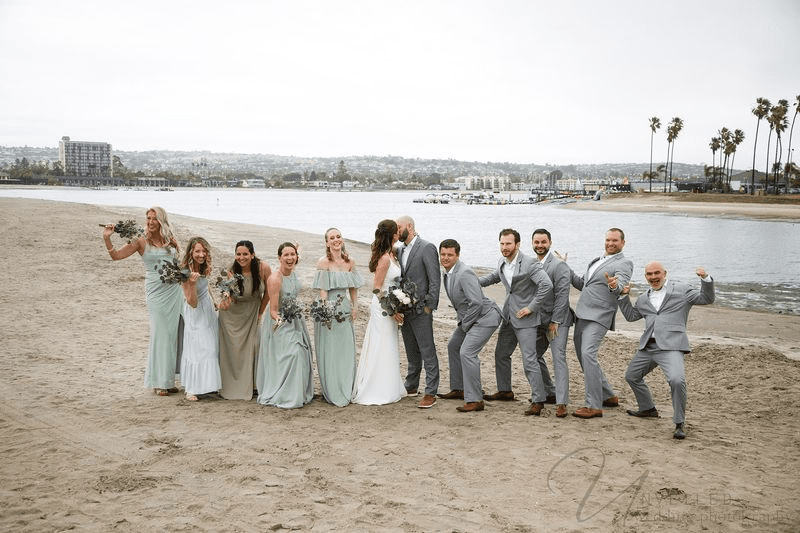 Bride and groom kissing while bridesmaids and groomsmen pose on the shore.