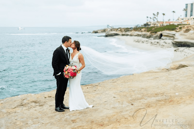 Bride and groom kissing on a rocky coastline with a long veil catching wind.