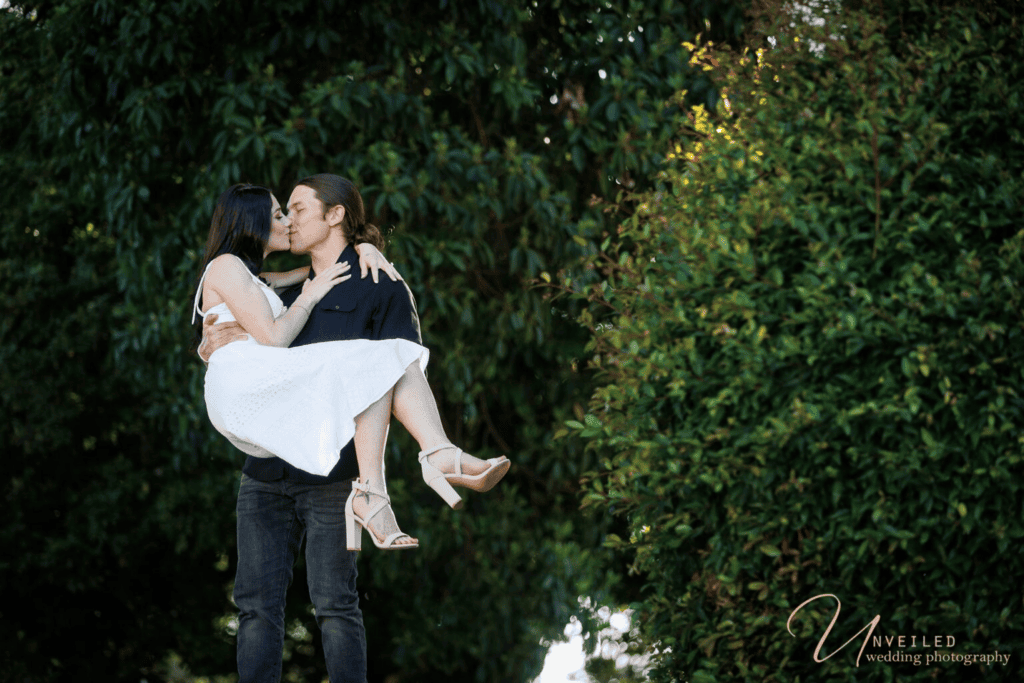 Man lifting and kissing a woman in a white dress during an outdoor photoshoot.