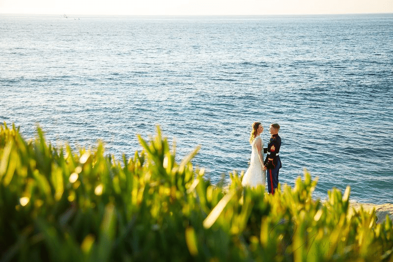A military groom holding hands with his bride on a cliff by the blue ocean.