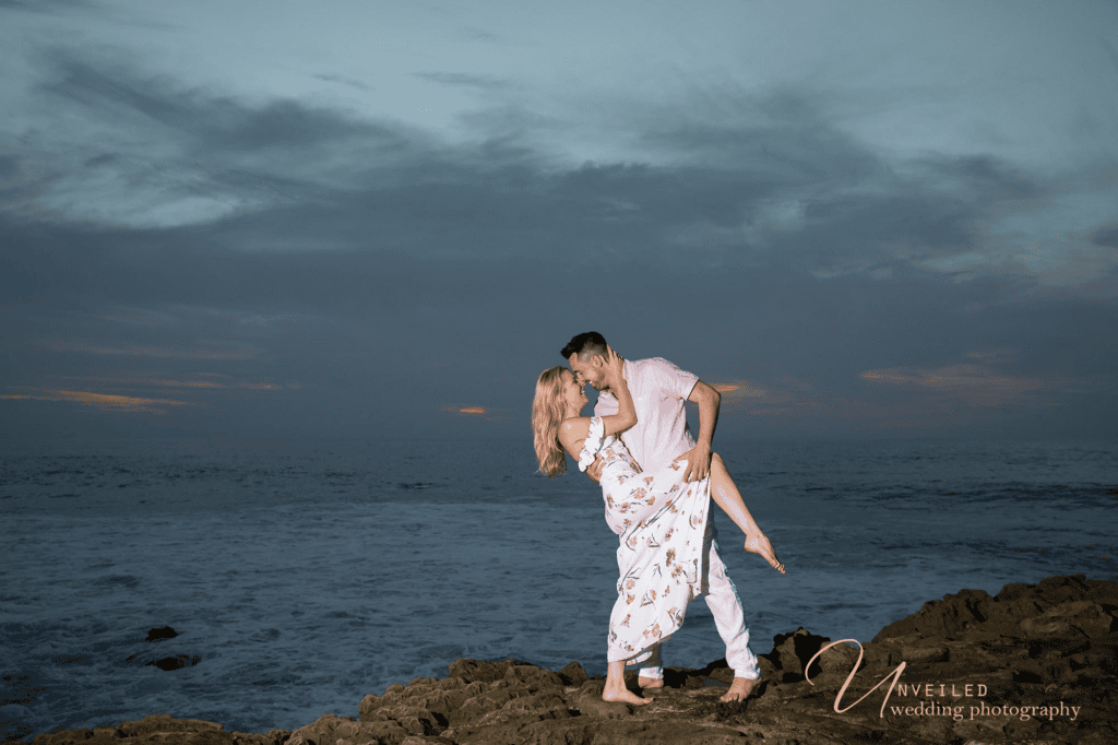 Groom lifting bride on rocky beach during an engagement session before your wedding.