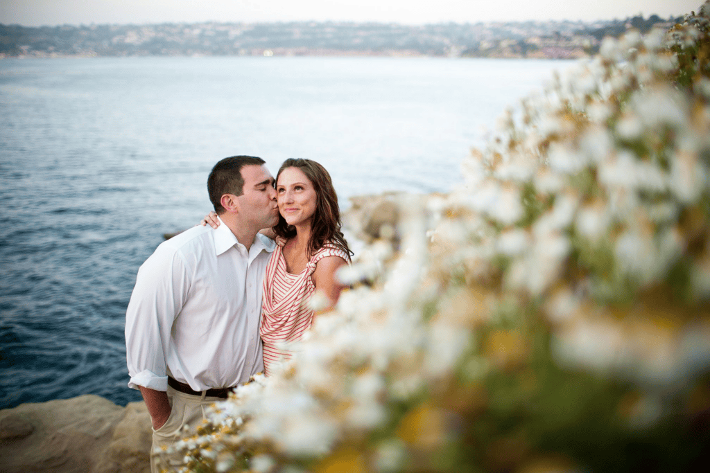 Man kissing woman on cheek near ocean flowers for engagement session before your wedding.