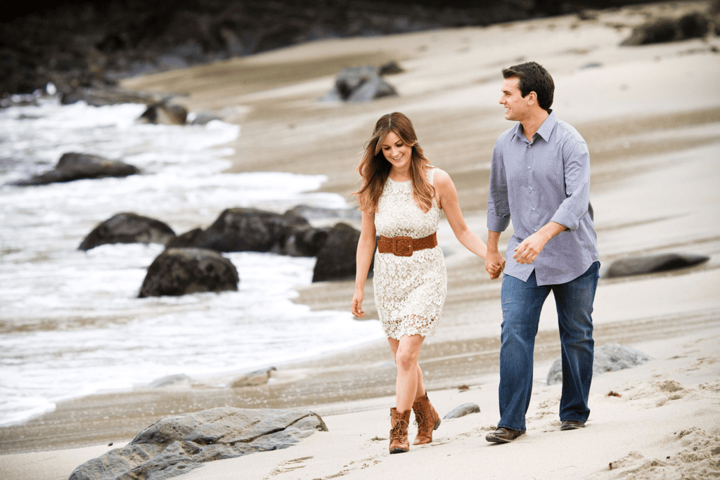 Couple walking on sandy beach holding hands for engagement session before your wedding.