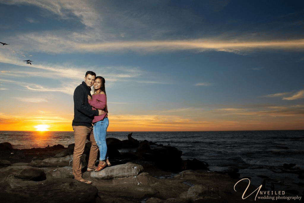 Couple posing on rocky shore at sunset by ocean waters for Engagement Photo Ideas.