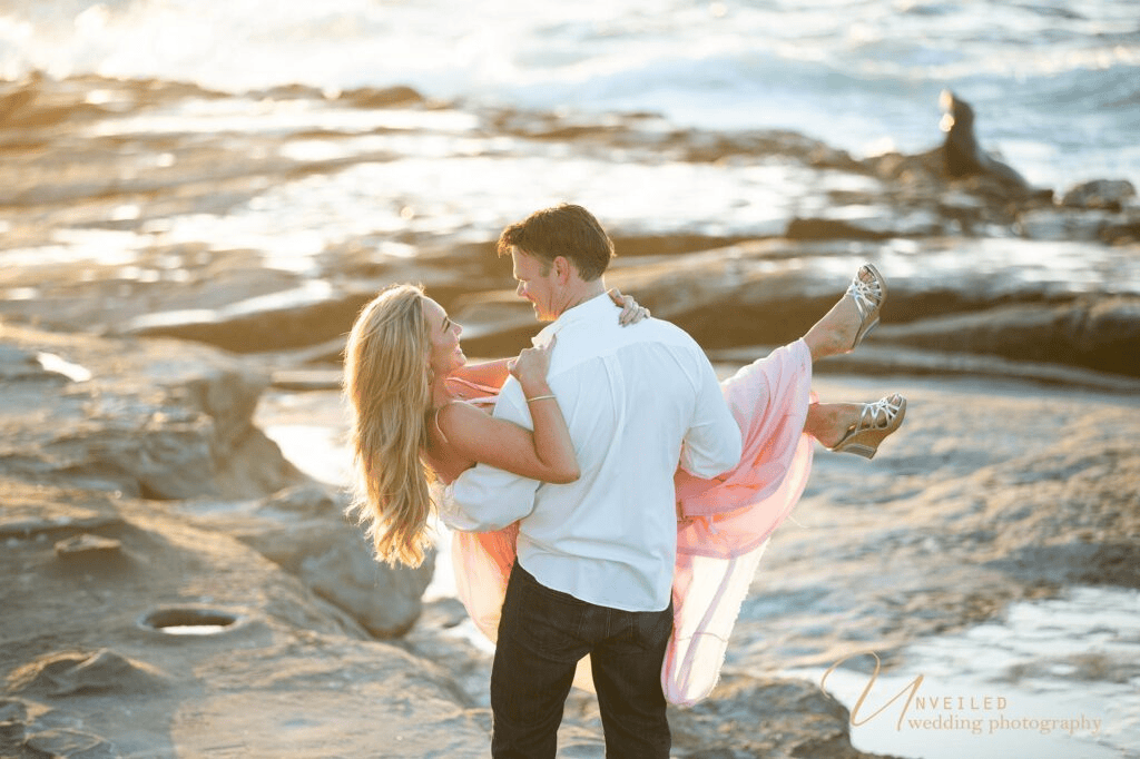 Groom holding bride in arms on rocky beach coast for Engagement Photo Ideas.
