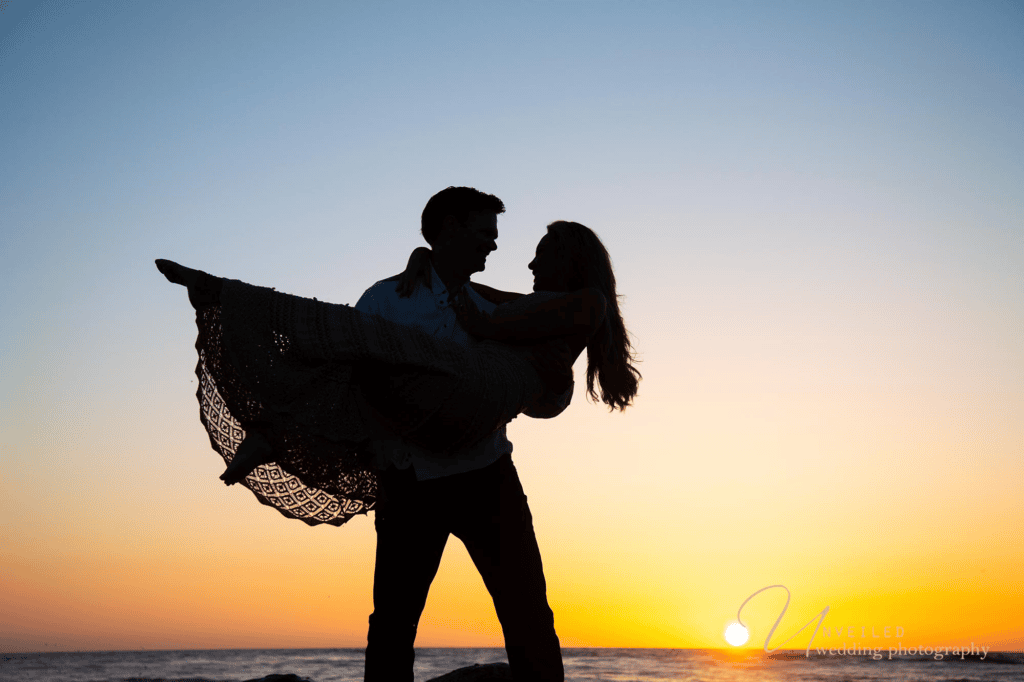 Sunset silhouette of man carrying fiancé on beach for dramatic Engagement Photo Ideas.