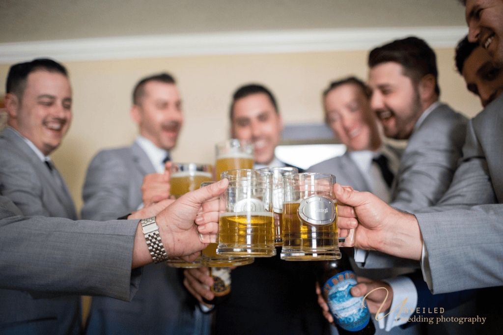 Groom and groomsmen toasting beer mugs by Grand Tradition Estate Wedding Photographer.